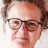 Porträt einer lächelnden Frau mit lockigem, hochgebundenem Haar. Sie trägt eine Brille und eine schlichte Bluse. Der Hintergrund ist hell und neutral, was den Fokus auf die Gesichtszüge und den freundlichen Ausdruck der Frau lenkt.