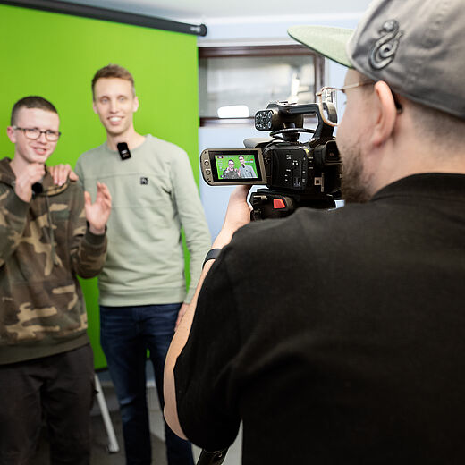 Zwei junge Männer stehen vor einem grünen Hintergrund und lächeln in die Kamera. Einer von ihnen hat eine Brille auf und trägt ein camouflages farbenes Oberteil, der andere trägt ein helles Sweatshirt. Im Vordergrund ist ein Kameramann zu sehen, der die Aufnahme macht.