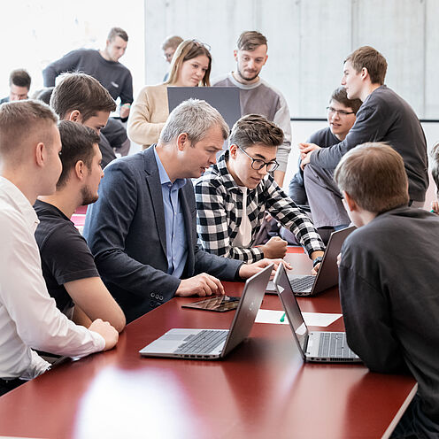 In einem modernen Büroraum arbeiten mehrere Menschen zusammen an Laptops. Eine Gruppe von fünf Personen, darunter ein älterer Mann und ein Jugendlicher, diskutiert aktiv und schaut auf einen Laptop. Im Hintergrund sind weitere Personen, die ebenfalls an ihren Geräten beschäftigt sind.
