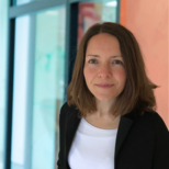 Eine Frau mit mittellangen, braunen Haaren trägt einen schwarzen Blazer und ein weißes T-Shirt. Sie schaut direkt in die Kamera und steht vor einem unscharfen, modernen Hintergrund.