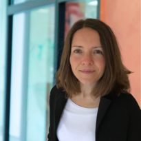 Eine Frau mit mittellangen, braunen Haaren trägt einen schwarzen Blazer und ein weißes T-Shirt. Sie schaut direkt in die Kamera und steht vor einem unscharfen, modernen Hintergrund.