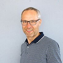 Ein Mann mit Brille lächelt freundlich. Er trägt ein gestreiftes Poloshirt und steht vor einer grauen Wand. Die Haltung wirkt entspannt und offen.