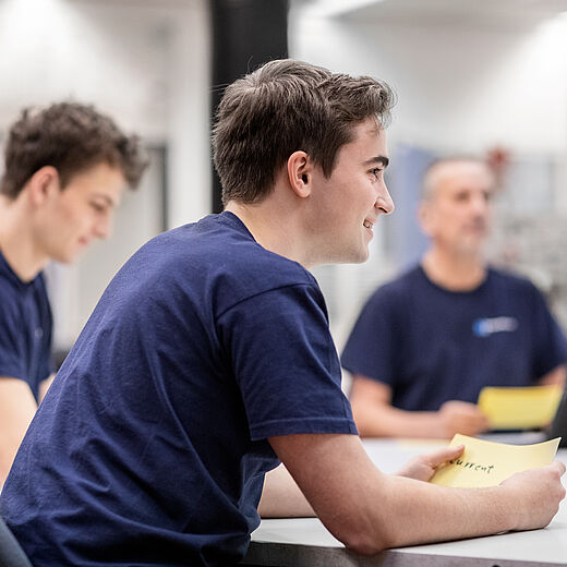 Drei Personen sitzen an einem Tisch in einem Innenraum. Zwei junge Männer tragen dunkelblaue T-Shirts und lächeln, während sie Notizen oder Karten in der Hand halten. Ein älterer Mann im Hintergrund wirkt aufmerksam, er verwendet ein Tablet. Die Atmosphäre ist konzentriert und freundlich.