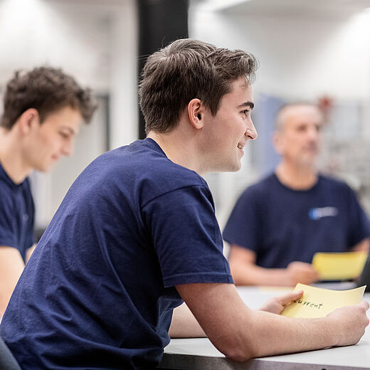 Drei Personen sitzen an einem Tisch in einem Innenraum. Zwei junge Männer tragen dunkelblaue T-Shirts und lächeln, während sie Notizen oder Karten in der Hand halten. Ein älterer Mann im Hintergrund wirkt aufmerksam, er verwendet ein Tablet. Die Atmosphäre ist konzentriert und freundlich.