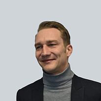 Ein Mann mit kurzen, hellen Haaren trägt einen grauen Rollkragenpullover und ein dunkles Jacket. Er steht leicht zur Seite und lächelt. Der Hintergrund ist einfarbig hellgrau.
