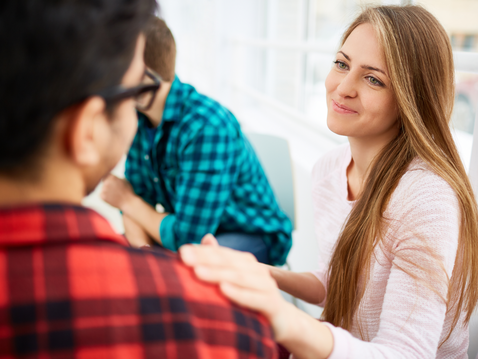 Eine Frau mit langen, blonden Haaren lächelt freundlich zu einem Mann, der ihr gegenüber sitzt. Sie legt eine Hand auf seine Schulter. Im Hintergrund sind zwei weitere Personen in einem hellen, modern eingerichteten Raum zu sehen.