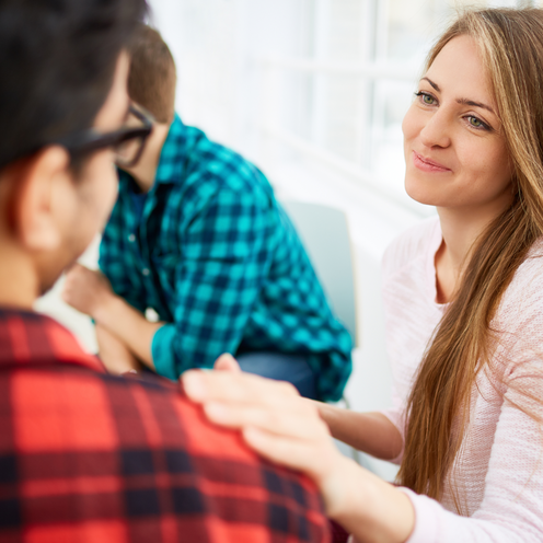 Eine Frau mit langen, blonden Haaren lächelt freundlich zu einem Mann, der ihr gegenüber sitzt. Sie legt eine Hand auf seine Schulter. Im Hintergrund sind zwei weitere Personen in einem hellen, modern eingerichteten Raum zu sehen.