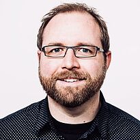 Ein lächelnder Mann mit Brille und einem Vollbart steht vor einem einfarbigen Hintergrund. Er trägt ein schwarzes Hemd mit einem auffälligen Muster und wirkt freundlich und zugänglich.