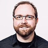 Ein lächelnder Mann mit Brille und einem Vollbart steht vor einem einfarbigen Hintergrund. Er trägt ein schwarzes Hemd mit einem auffälligen Muster und wirkt freundlich und zugänglich.