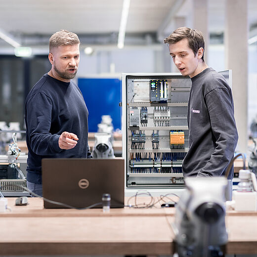 Zwei Personen in einem technischen Arbeitsumfeld diskutieren an einem Tisch. Einer zeigt auf ein offenes Steuerungssystem, während der andere aufmerksam zuhört. Im Hintergrund sind weitere technische Geräte sichtbar.