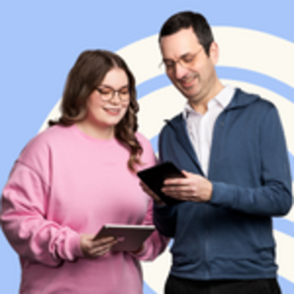 Zwei Personen stehen nebeneinander und schauen gemeinsam auf digitale Geräte. Eine Person trägt einen pinken Pullover, die andere einen blauen. Beide wirken konzentriert und tauschen sich über das, was sie sehen. Der Hintergrund ist hellblau mit einem dekorativen Muster.