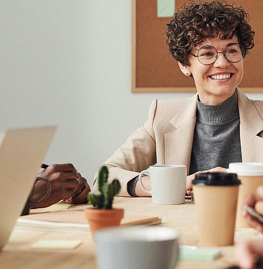 Eine Gruppe von fünf Personen sitzt an einem Tisch in einem Besprechungsraum. Die Atmosphäre ist freundlich und kollegial, während sie miteinander kommunizieren. Auf dem Tisch stehen Kaffee-Becher und ein Laptop ist geöffnet.