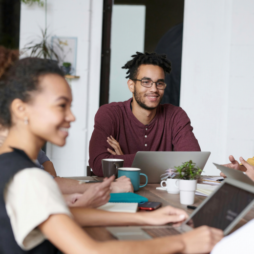 Eine Gruppe von fünf Menschen sitzt an einem Tisch, der mit Laptops, Notizblöcken und Kaffeetassen gedeckt ist. Sie lächeln und scheinen an einem kreativen Austausch oder einem Meeting beteiligt zu sein. Der Raum hat große Fenster, die natürliches Licht hereinlassen.
