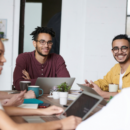 Eine Gruppe von fünf Menschen sitzt an einem Tisch, der mit Laptops, Notizblöcken und Kaffeetassen gedeckt ist. Sie lächeln und scheinen an einem kreativen Austausch oder einem Meeting beteiligt zu sein. Der Raum hat große Fenster, die natürliches Licht hereinlassen.