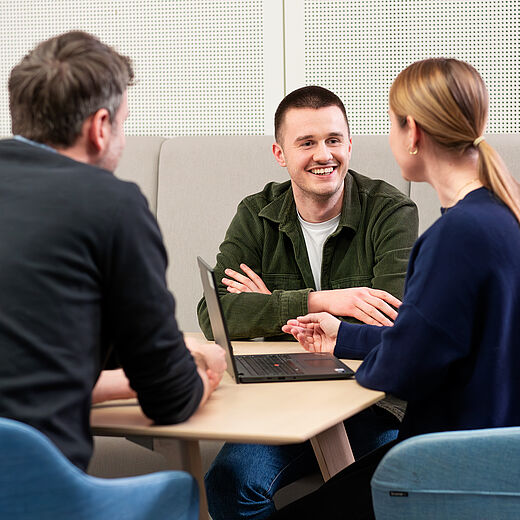 Drei Personen sitzen an einem Tisch in einem modernen Besprechungsraum. Sie diskutieren lebhaft, während ein Laptop vor ihnen steht. Eine Person lächelt, während die anderen aufmerksam zuhören. Die Atmosphäre wirkt freundlich und kooperativ.