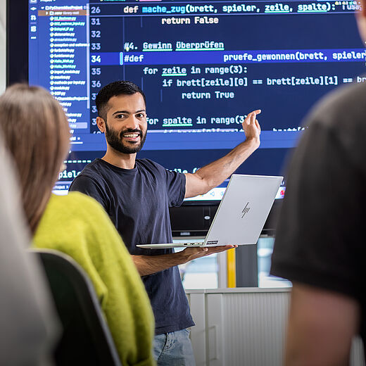 Ein Mann mit dunklen Haaren hält einen Laptop in der Hand und erklärt etwas vor einer großen Bildschirmpräsentation mit Programmiercode. Im Hintergrund sind einige Personen zu sehen, die aufmerksam zuhören. Der Mann schaut lächelnd in die Runde.