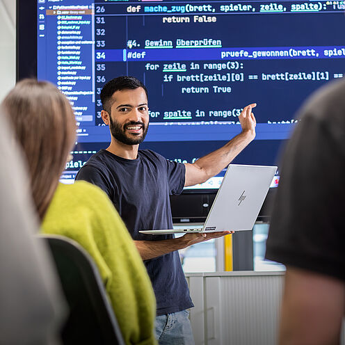 Ein Mann mit dunklen Haaren hält einen Laptop in der Hand und erklärt etwas vor einer großen Bildschirmpräsentation mit Programmiercode. Im Hintergrund sind einige Personen zu sehen, die aufmerksam zuhören. Der Mann schaut lächelnd in die Runde.
