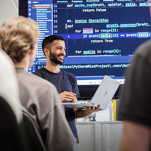Ein lächelnder Dozent steht mit einem Laptop in der Hand vor einer Gruppe von Menschen. Im Hintergrund sind Programmierzeilen auf einem Bildschirm zu sehen. Die Teilnehmer hören aufmerksam zu, während der Dozent etwas erklärt.