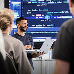 Ein lächelnder Dozent steht mit einem Laptop in der Hand vor einer Gruppe von Menschen. Im Hintergrund sind Programmierzeilen auf einem Bildschirm zu sehen. Die Teilnehmer hören aufmerksam zu, während der Dozent etwas erklärt.