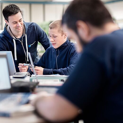 Zwei junge Männer sitzen an einem Tisch, einer hilft dem anderen mit einem technischen Gerät, während sie freundlich miteinander sprechen. Ein dritter Mann im Hintergrund ist mit einem Laptop beschäftigt. Die Szene vermittelt eine Atmosphäre der Zusammenarbeit und Unterstützung.