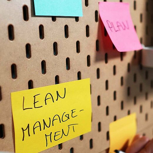Ein handgeschriebener Zettel mit der Aufschrift „LEAN MANAGEMENT“ ist auf einer Wand aus Lochplatten befestigt. Daneben sind weitere bunte Haftnotizen angebracht. Im Hintergrund ist ein Gerät zu sehen, das nicht näher beschrieben ist.