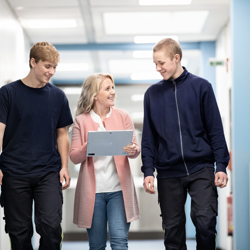 Drei Menschen gehen lächelnd einen Flur entlang. Zwei junge Männer in Arbeitskleidung stehen neben einer Frau, die ein Tablet hält. Die Atmosphäre wirkt freundlich und informell, als ob sie sich über etwas Wichtiges austauschen.