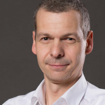 Porträt eines Mannes mit kurzen, grauen Haaren und einem neutralen Ausdruck. Er trägt ein weißes Hemd und steht vor einem dunklen Hintergrund.