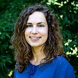 Ein Porträt einer jungen Frau mit lockigem, braunem Haar, die in die Kamera lächelt. Sie trägt eine blaue Bluse. Im Hintergrund sind grüne Pflanzen sichtbar, die eine natürliche Umgebung schaffen.