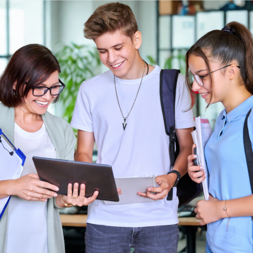 Eine Frau zeigt zwei lächelnden Jugendlichen in einem hellen Büro oder Klassenzimmer etwas auf einem Tablet, während alle gemeinsam darauf schauen.