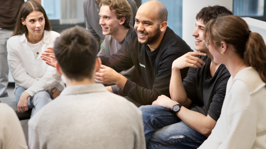 Eine Gruppe von sechs Personen sitzt in einem offenen Raum und lacht miteinander. Einige von ihnen sprechen und gestikulieren, während andere aufmerksam zuhören. Die Atmosphäre wirkt freundlich und kommunikativ, was ein Gefühl von gemeinsamen Austausch und Teamarbeit vermittelt.
