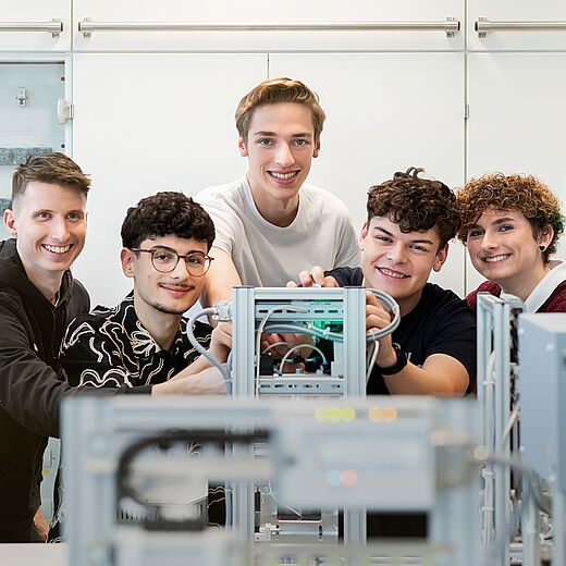 Fünf junge Menschen stehen in einem technischen Raum und arbeiten gemeinsam an einem Gerät. Sie lächeln und scheinen Freude an der Zusammenarbeit zu haben. Im Hintergrund sind technische Geräte zu sehen.