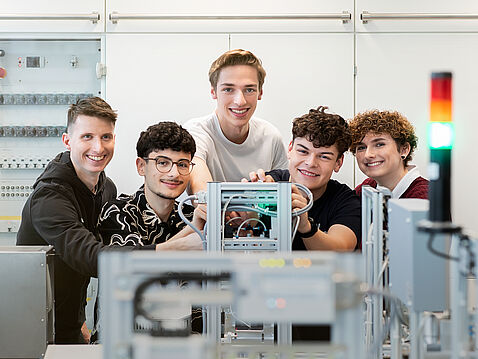Fünf junge Menschen stehen in einem technischen Raum und arbeiten gemeinsam an einem Gerät. Sie lächeln und scheinen Freude an der Zusammenarbeit zu haben. Im Hintergrund sind technische Geräte zu sehen.