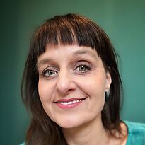 Porträt einer Frau mit langen, braunen Haaren und einem freundlichen Lächeln. Sie trägt ein türkisfarbenes Oberteil und hat schlichte Ohrringe. Der Hintergrund ist grün. Die Frau schaut direkt in die Kamera, was eine einladende und positive Ausstrahlung vermittelt.