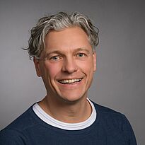 Porträt eines lächelnden Mannes mit lockigen, hellen Haaren. Er trägt ein dunkelblaues Oberteil mit weißem Rundhalsausschnitt. Der Hintergrund ist grau und neutral. Seine Ausstrahlung ist freundlich und offen.