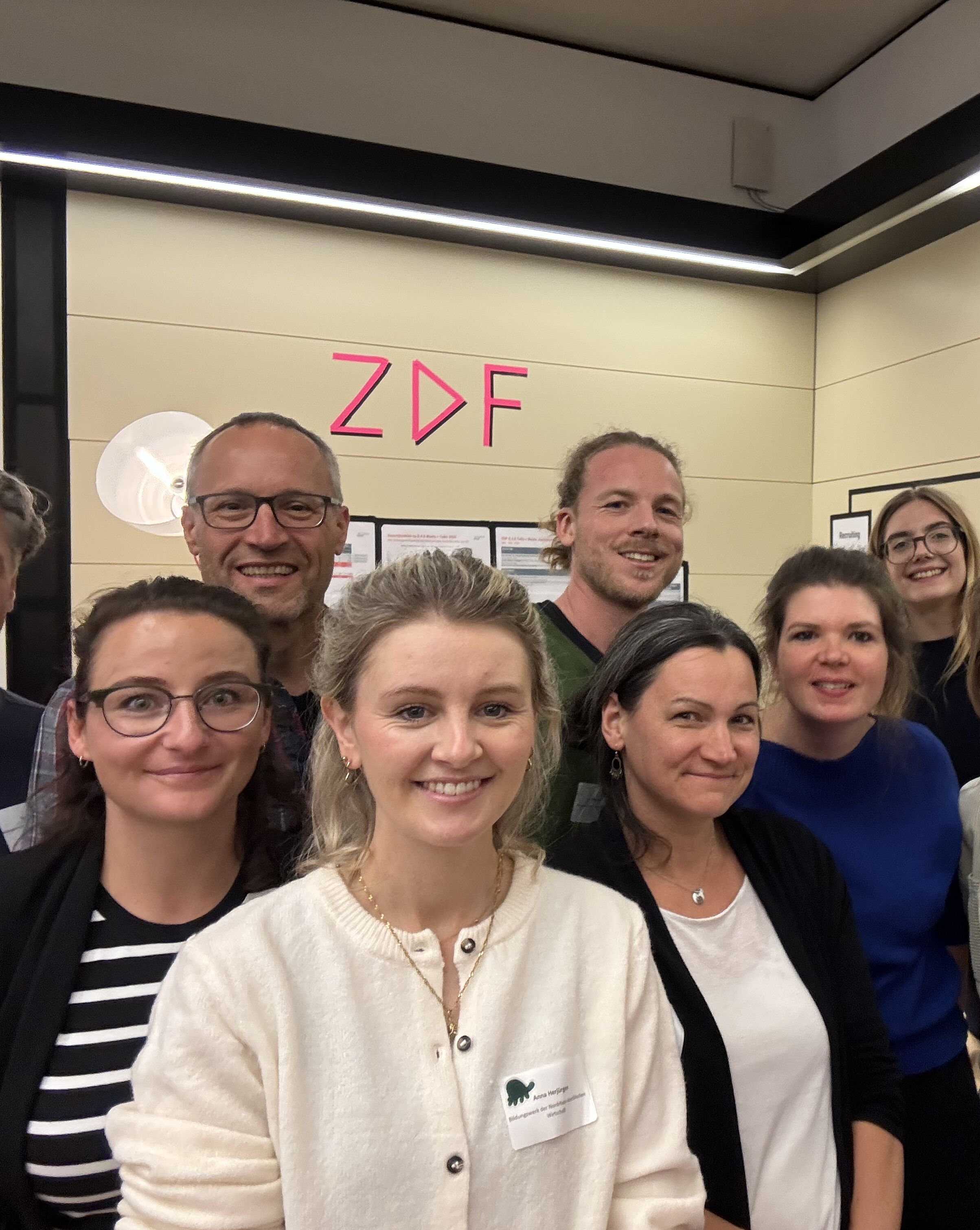 Eine Gruppe von zehn Personen steht in einem Raum zusammen und lächelt freundlich in die Kamera. Im Hintergrund sind Wandtafeln sichtbar. Die Atmosphäre wirkt freundlich und entspannt, der Raum scheint hell beleuchtet zu sein.