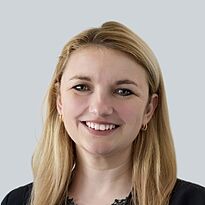 Eine junge Frau mit langen, blonden Haaren lächelt freundlich in die Kamera. Sie trägt eine schwarze Bluse und hat goldene Ohrringe. Der Hintergrund ist einfarbig grau.