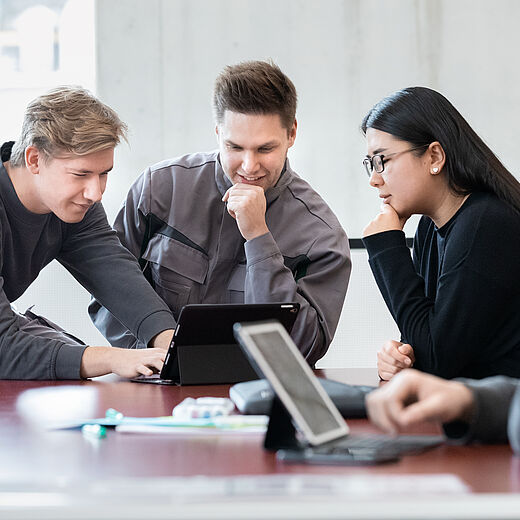 Vier Personen sitzen an einem Tisch und betrachten gemeinsam ein Tablet. Zwei Männer und eine Frau scheinen konzentriert zu diskutieren, während ein weiterer Mann am Tisch etwas abseits arbeitet. Der Raum hat große Fenster und eine moderne Einrichtung.