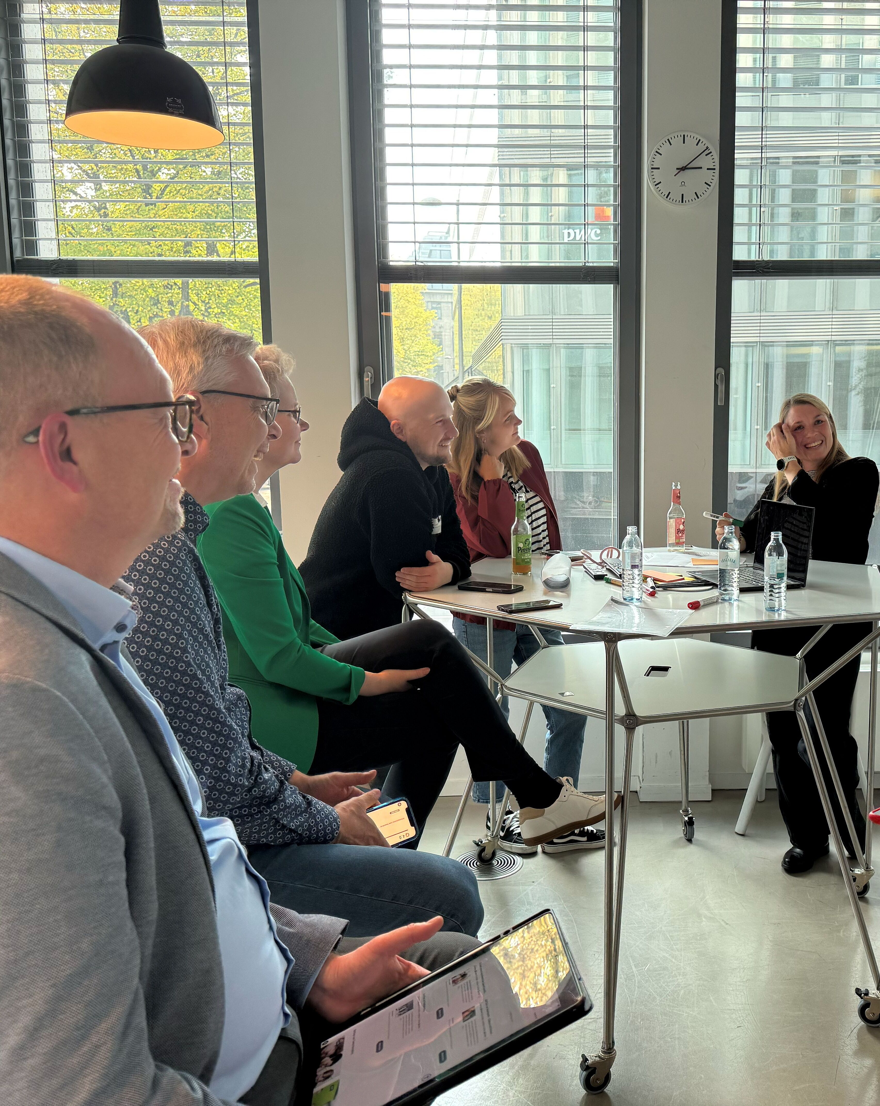 Eine Gruppe von sechs Personen sitzt an einem Tisch in einem modernen Konferenzraum. Einige Personen diskutieren, während andere zuhören. Der Raum hat große Fenster mit Blick nach draußen und ist mit Flipcharts und Wasserflaschen ausgestattet. An der Wand hängt eine Uhr.