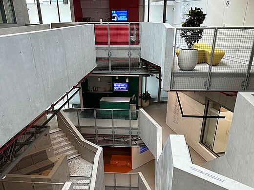Ein Blick in ein modernes, mehrstöckiges Gebäude. Sichtbar sind helle Treppen und verschiedene farbige Flächen auf den Etagen. Eine Stahlgeländer ist vorhanden, und im Hintergrund stehen einige Pflanzen und Möbel. Das Design ist funktional und ansprechend.